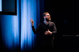 A bearded UX london speaker gestures to the screen behind him.